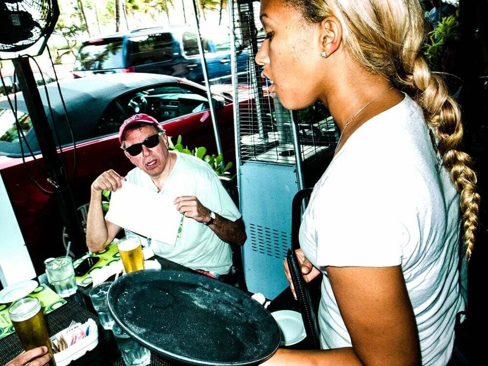 Colour flash photo of a waitress with a braid carrying a tray past a man at a restaurant table