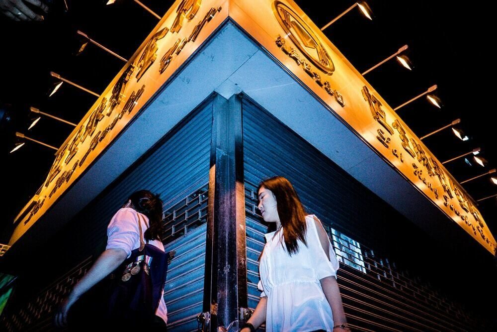 Colour flash photo shot from below of two women under a glowing orange sign against a blue sky