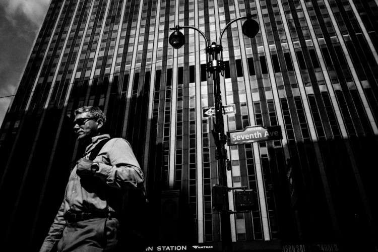 Black and white street photo of a man walking past a tall striped building, framed by a large black shadow on the building behind him