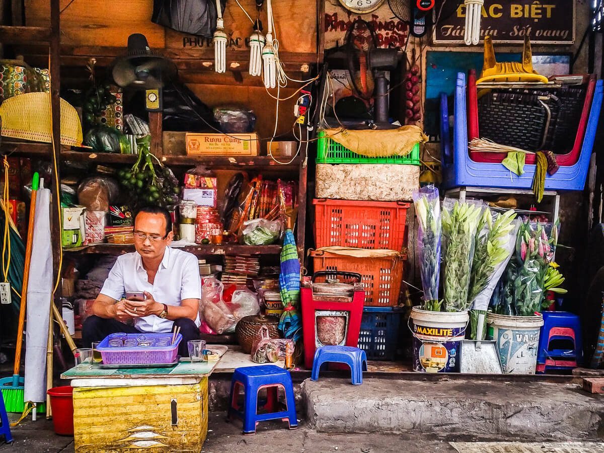 Color photo of a Hanoi shopkeeper sitting inside a small storefront full of goods, framed by the shop's edges