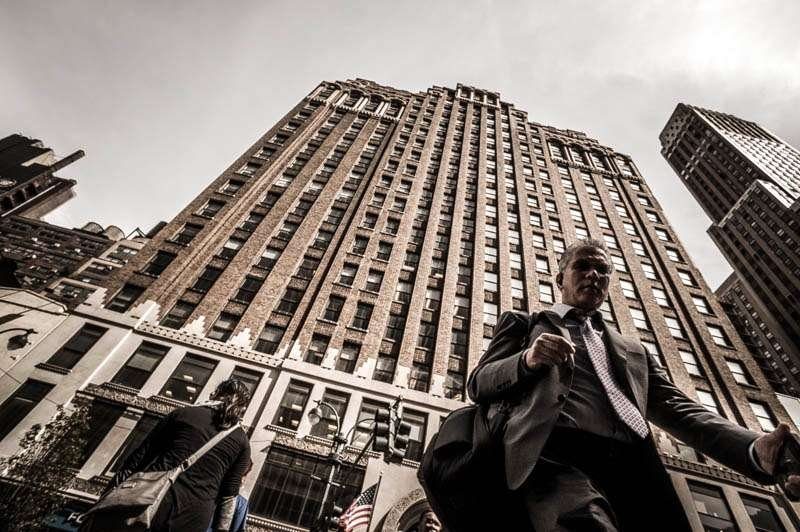 Businessman in a suit walking past a tall art deco building, shot from a low angle with a wide lens so the building leans