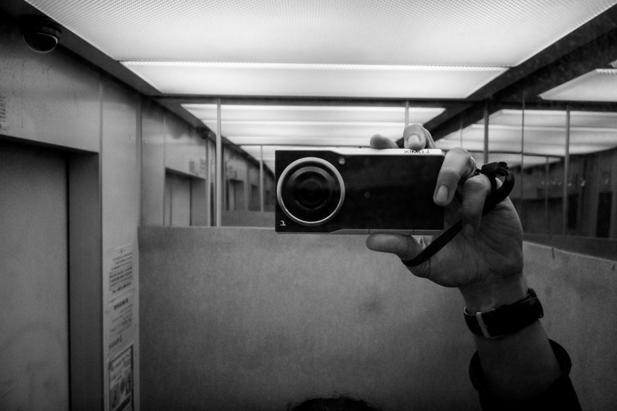 A hand holding a small camera up to a reflective wall in a corridor, black and white