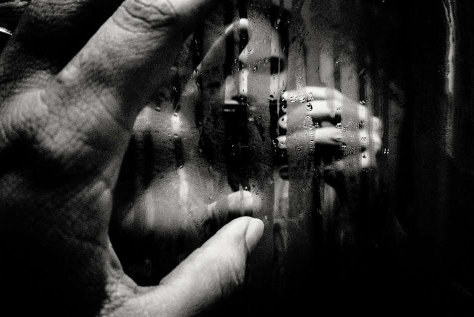 Close-up of a hand pressed against rain-streaked glass with a figure blurred behind, black and white