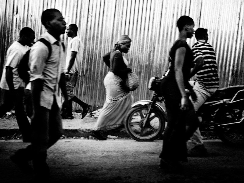 People walking past a corrugated metal fence on a busy street, black and white