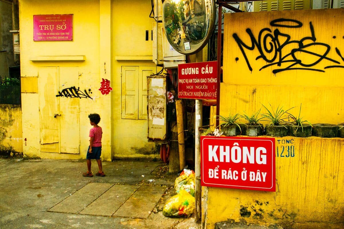Small child in a red shirt standing alone in front of a bright yellow wall with signs and graffiti in Hanoi