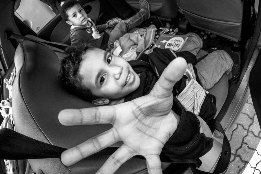 Black and white wide angle photo of a laughing boy reaching his open hand toward the camera from the back seat of a car