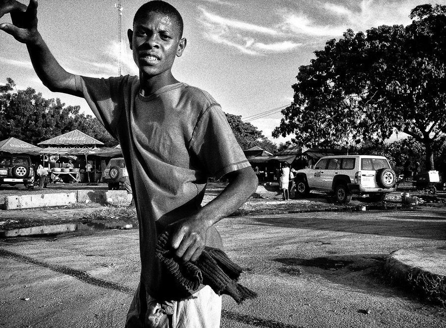 Black and white photo of a young man raising one arm in an open lot, with trees and a truck behind him and a dramatic sky