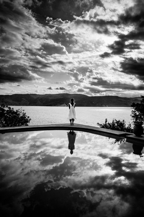 A small figure in a white dress standing on the edge of a pool that reflects a dramatic cloudy sky