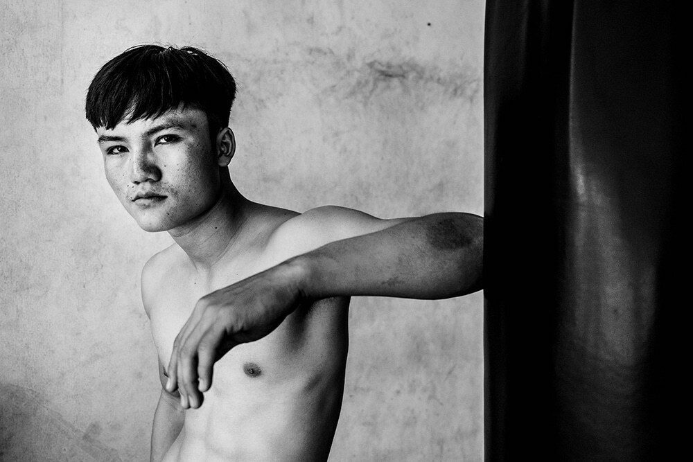 Posed black and white street portrait of a shirtless young man leaning on a wall, looking at the camera