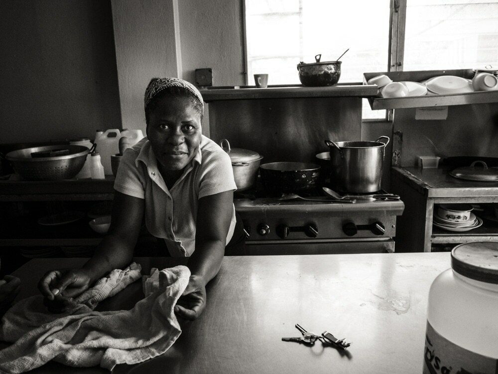 Black and white environmental portrait of a woman wiping a counter in a Haitian kitchen, with pots and a stove behind her