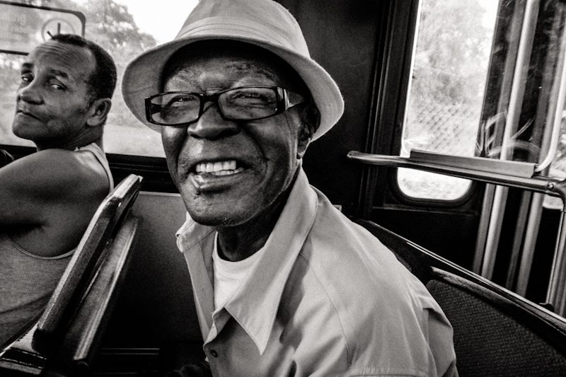 Smiling older man in a hat on a bus, another passenger glancing at the camera