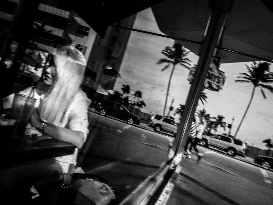 Woman reflected in a café window with palm trees and a street outside, black and white