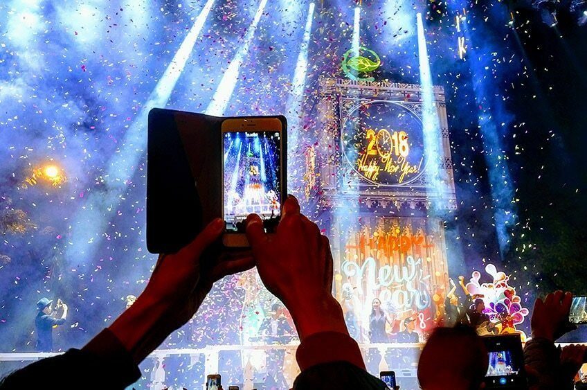 A hand holds up a phone to film a New Year's countdown show, with blue light beams and confetti in the background