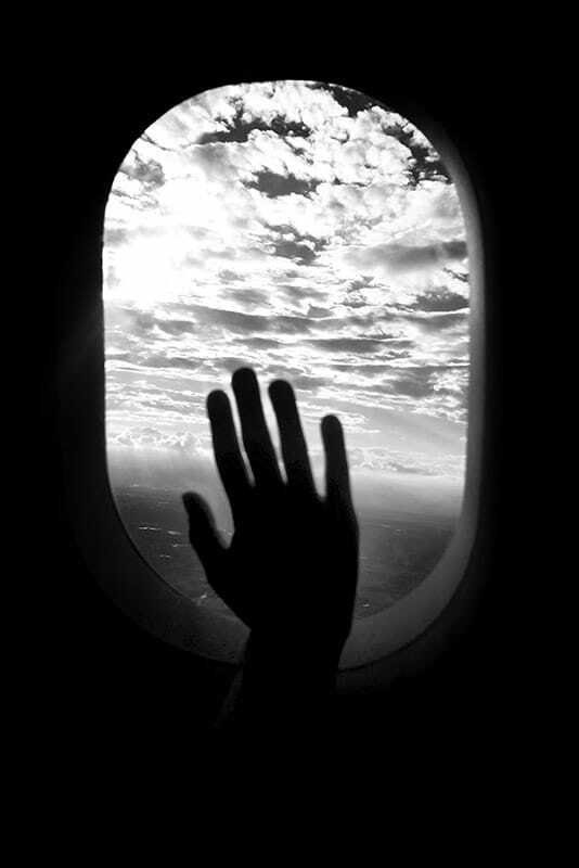Black and white photo of a hand pressed against an airplane window with clouds outside