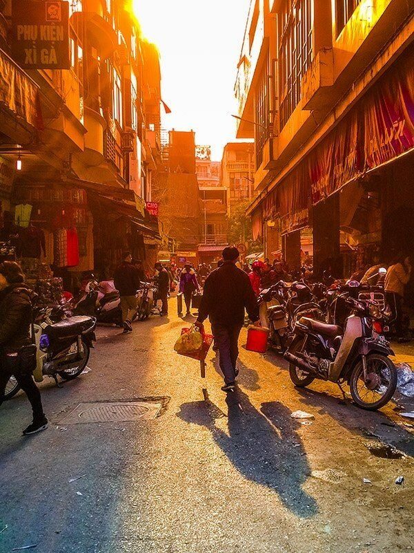 A busy narrow street at sunset, with low golden light and people and motorbikes in silhouette