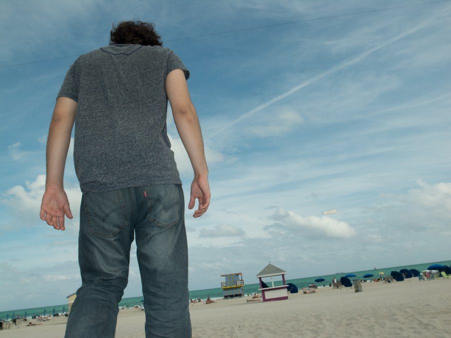 Color photo taken from a very low angle of a man in jeans standing on a beach under a blue sky with lifeguard huts behind him