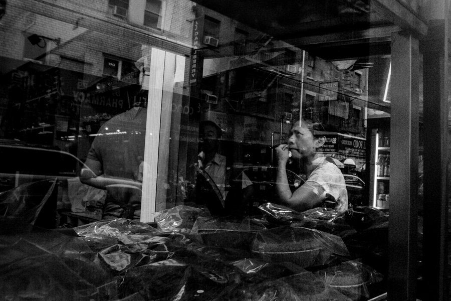 Black and white photo through a shop window, a man eating at a counter behind reflections and objects in the glass