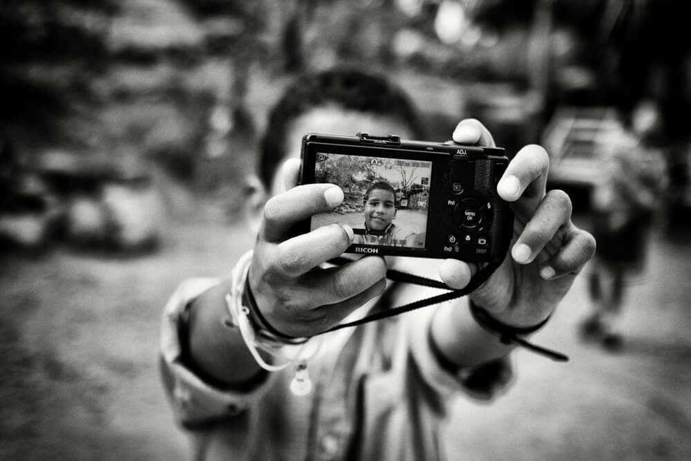 Black and white photo of a smiling boy holding up a small camera toward the lens, showing his own picture on the screen