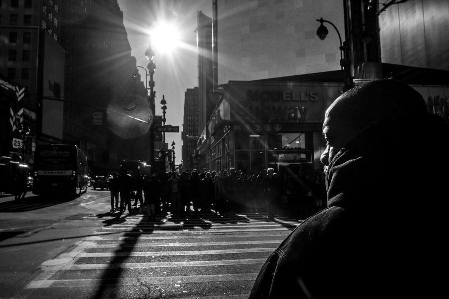 Black and white street photo of a crosswalk with the sun low between buildings and a hooded figure in the foreground