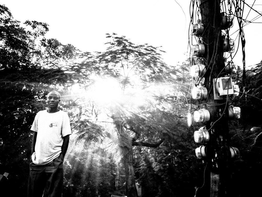 Black and white photo of a man standing under trees with sunbeams flaring through the leaves behind him and a pole of electric meters on the right