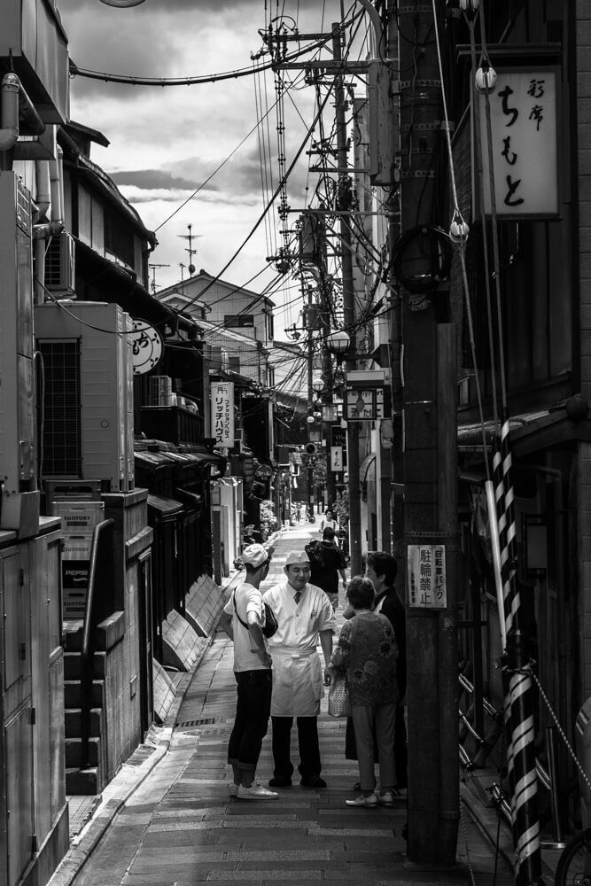 Black and white photo of a narrow Kyoto lane with a few people standing and talking in the distance