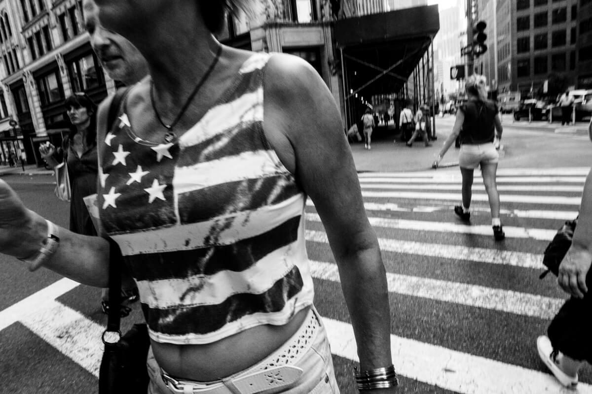Black and white photo of a woman in a stars and stripes top crossing a busy crosswalk