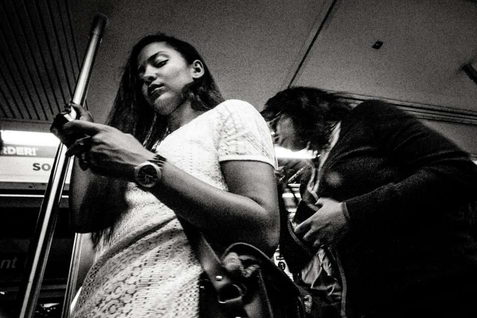 Black and white photo of a young woman checking her phone on a subway train, another rider beside her