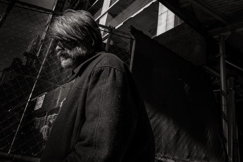 Black and white photo of a long haired older man in a heavy coat beside a chain link fence
