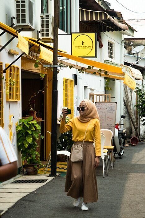 Woman in a yellow top and brown skirt photographing with a phone in a narrow street with yellow awnings in Penang