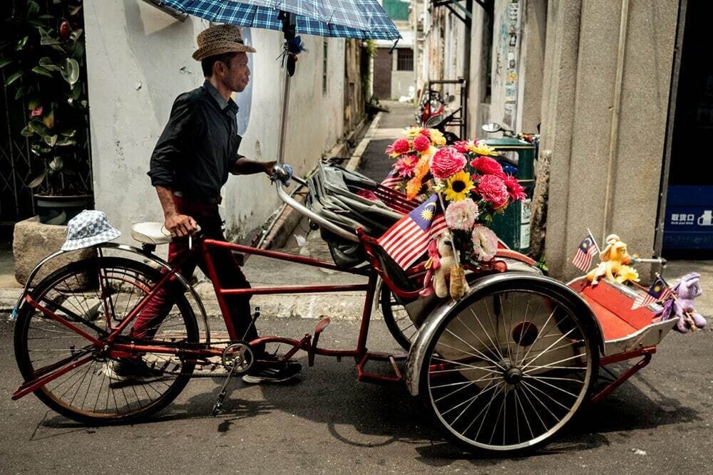 Man pushing a trishaw decorated with bright flowers and a small flag on a Penang street