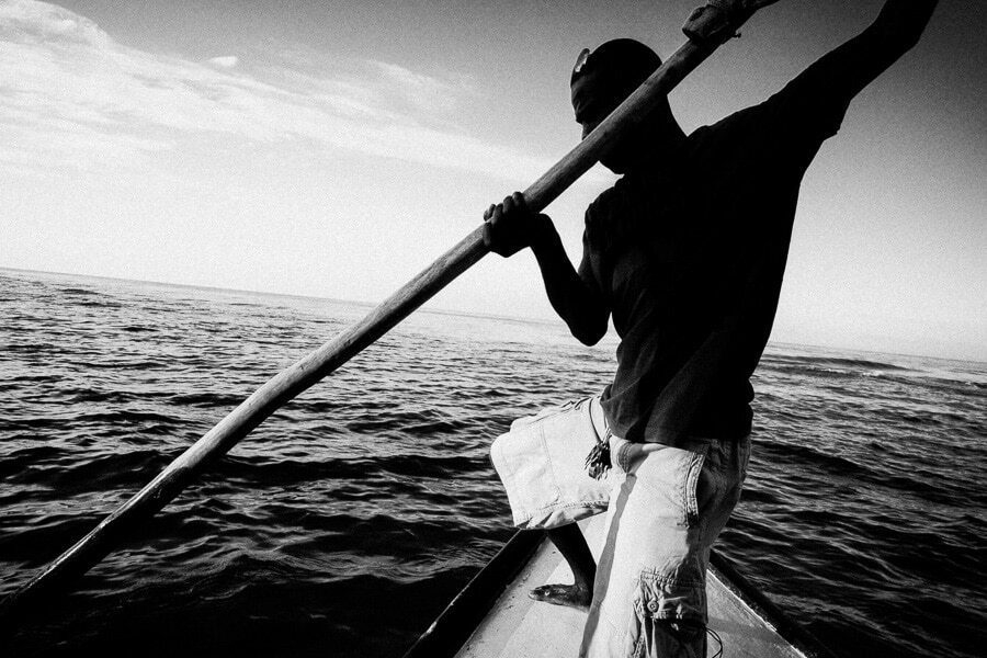 Black and white photograph of a man pushing a long pole against the sea from a boat, shot close with a wide angle