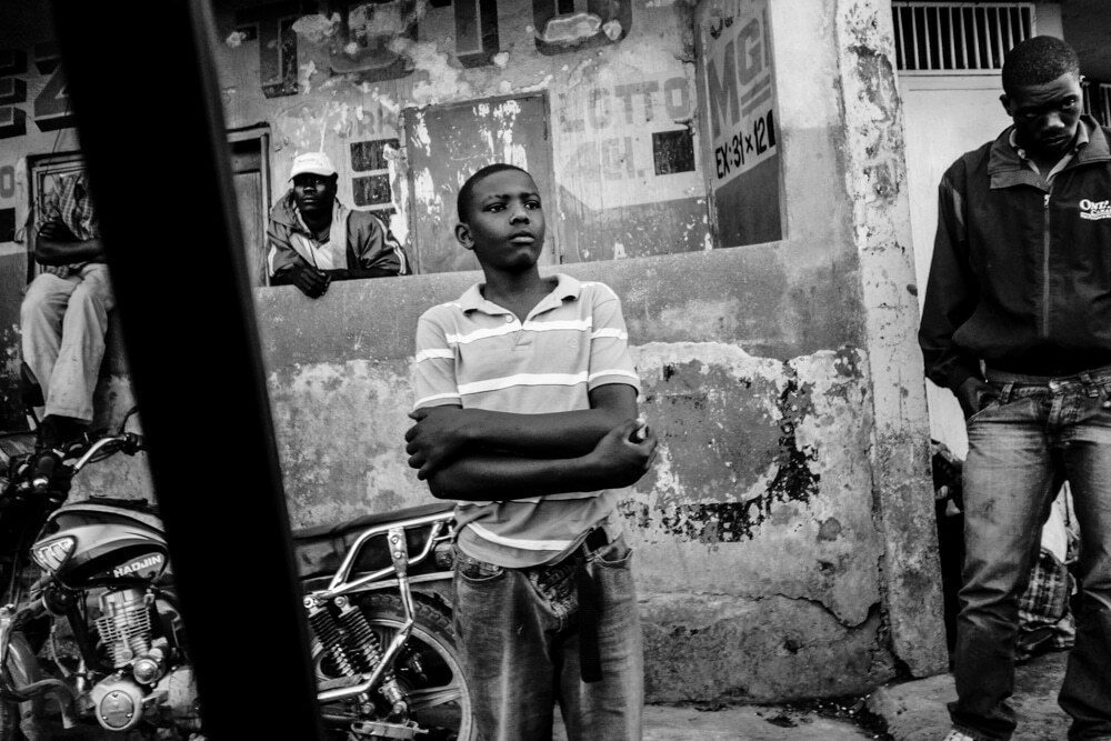 Black and white street scene in Haiti with a dark post in the foreground, a boy with crossed arms in the middle and men by a wall behind