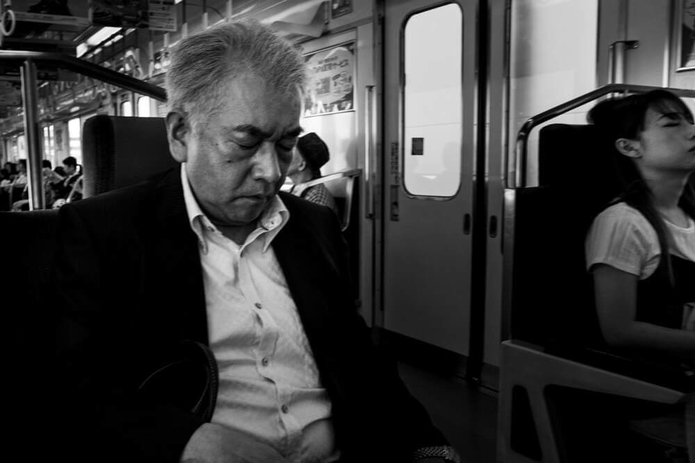 Man asleep on an Osaka train in the foreground with a young woman seated farther back