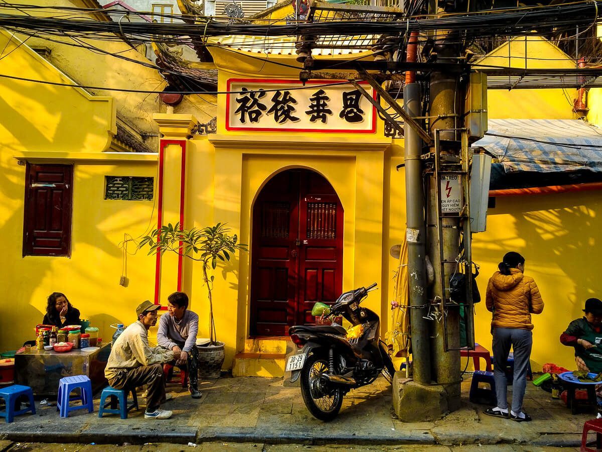 Hanoi street scene against a flat yellow wall with people sitting on small stools