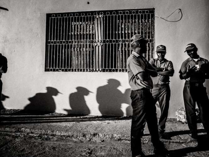 Black and white photo of three men standing by a white wall, with their shadows forming a line across the wall toward them