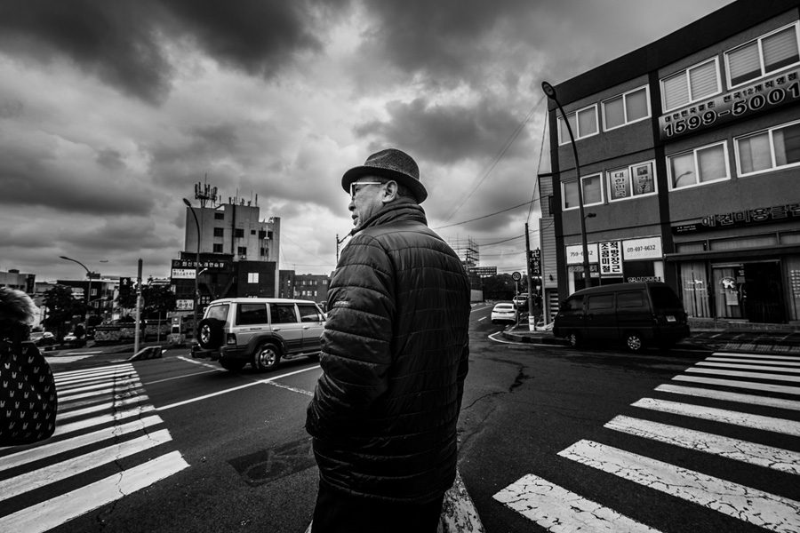 Wide angle black and white photo of a man in a hat standing at an intersection, with crosswalk stripes running diagonally toward him