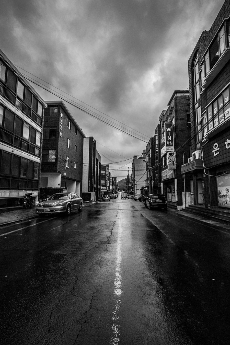 Vertical black and white photo of a wet city street shot from low, with the center line running straight to a vanishing point between buildings