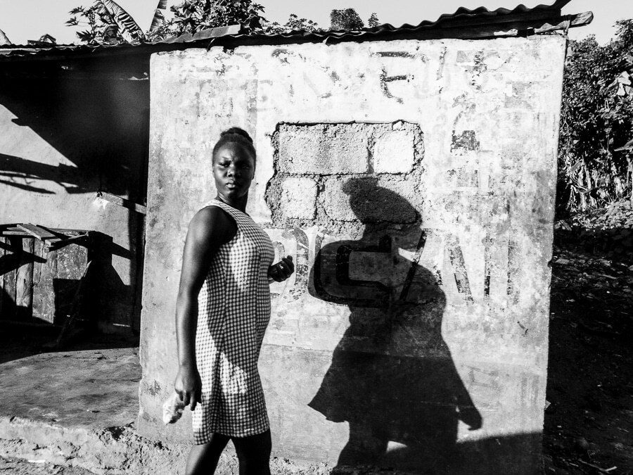 Black and white photo of a woman in a checked dress walking past a white wall in Haiti, with her dark shadow cast on the wall beside her