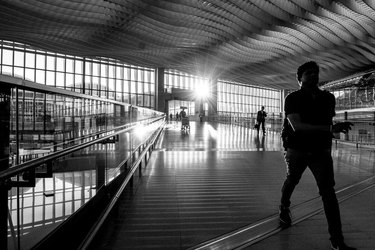 Black and white photo of a man walking through a Hong Kong airport hallway, backlit by the sun, with light reflecting on the shiny floor