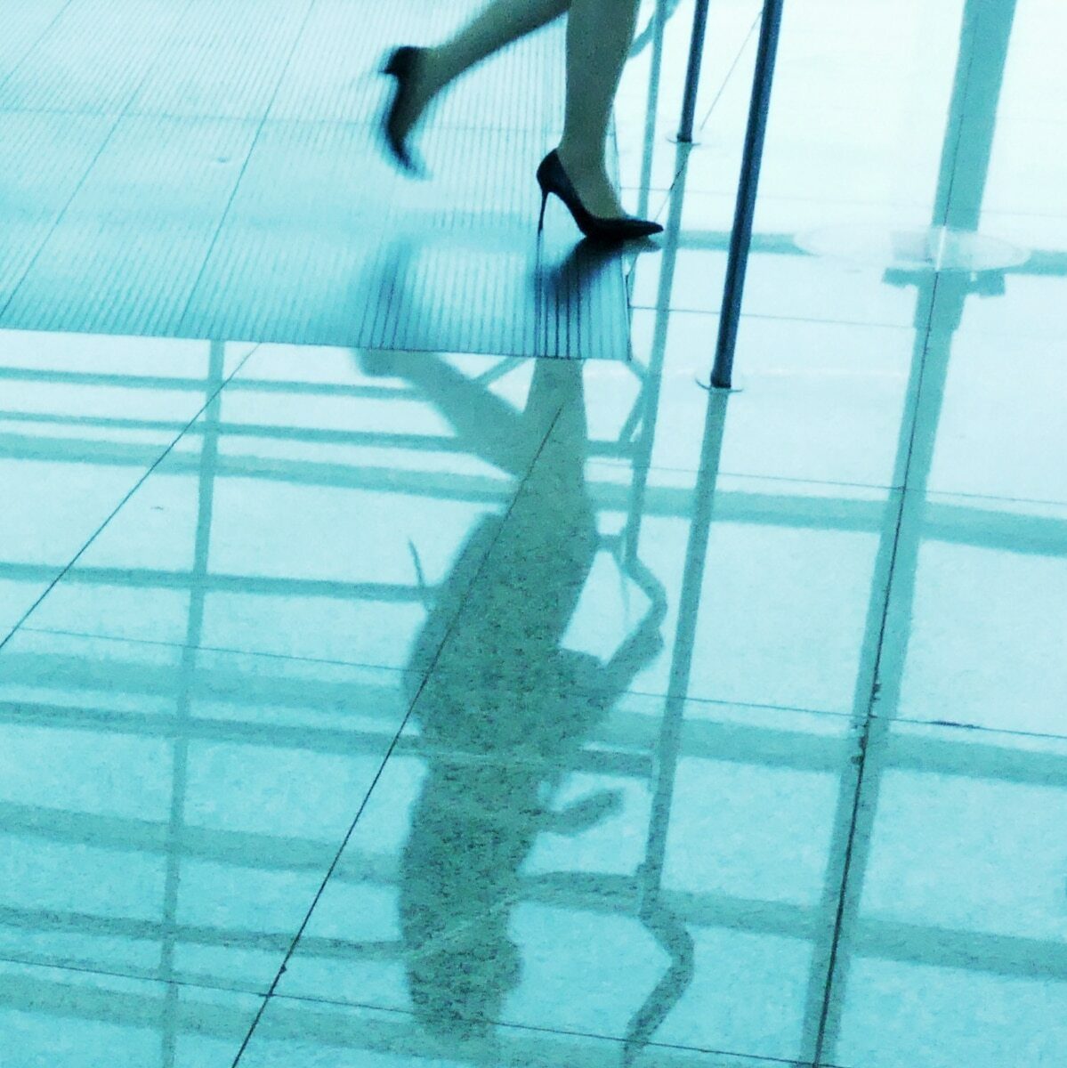 Blue tinted photo of a woman's legs in high heels walking across a glossy airport floor, her full reflection visible below