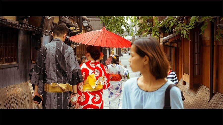 Tourists in rented kimonos and a woman with a red paper umbrella walking along a narrow old street in Kyoto