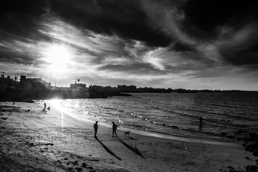 Black and white beach at sunset with small silhouetted figures walking along the shore under dramatic clouds