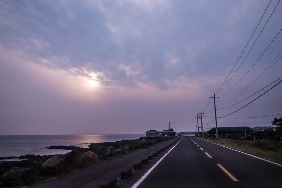 Empty coastal road at dusk under a purple sky with power lines and a low sun