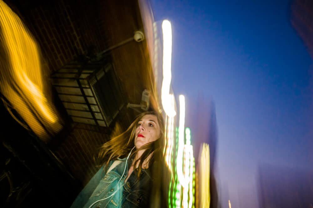 Colour photo of a woman in an evening street, face sharp, with neon lights streaking around her