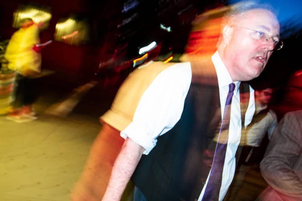 Colour flash photo of an older man in a vest and tie, sharp in the middle with coloured light streaks around him