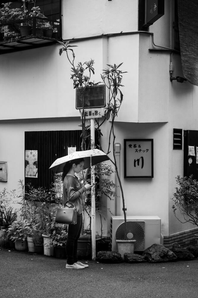 Black and white photo of a woman with a parasol standing beside a pale building in Kyoto, small in the frame