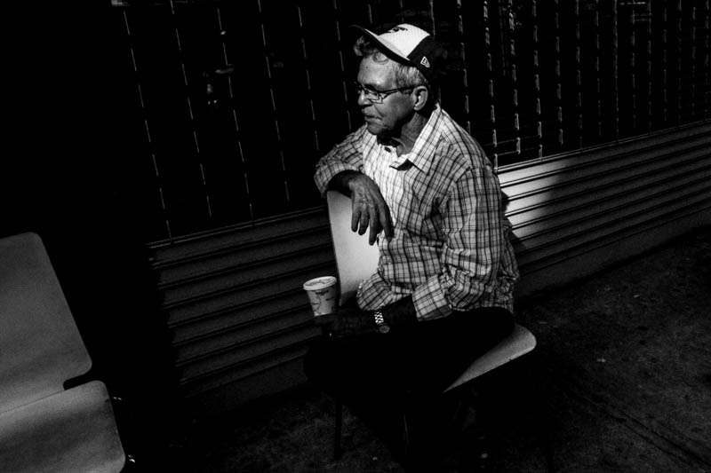 Black and white photo of an old man in a cap sitting in a patch of light surrounded by deep shadow