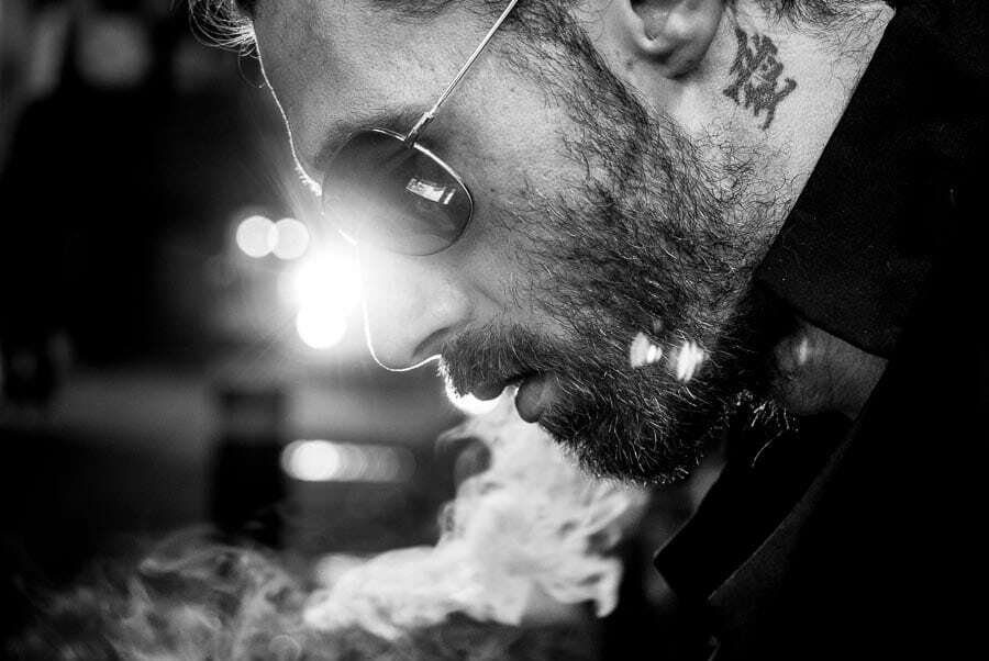 Black and white night portrait of a bearded man with sunglasses exhaling smoke, lit by a storefront