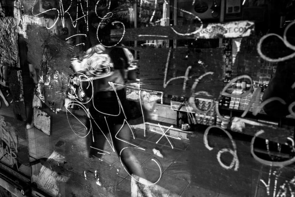 Black and white night photo through a shop window covered in scribbles, with reflections and a figure inside