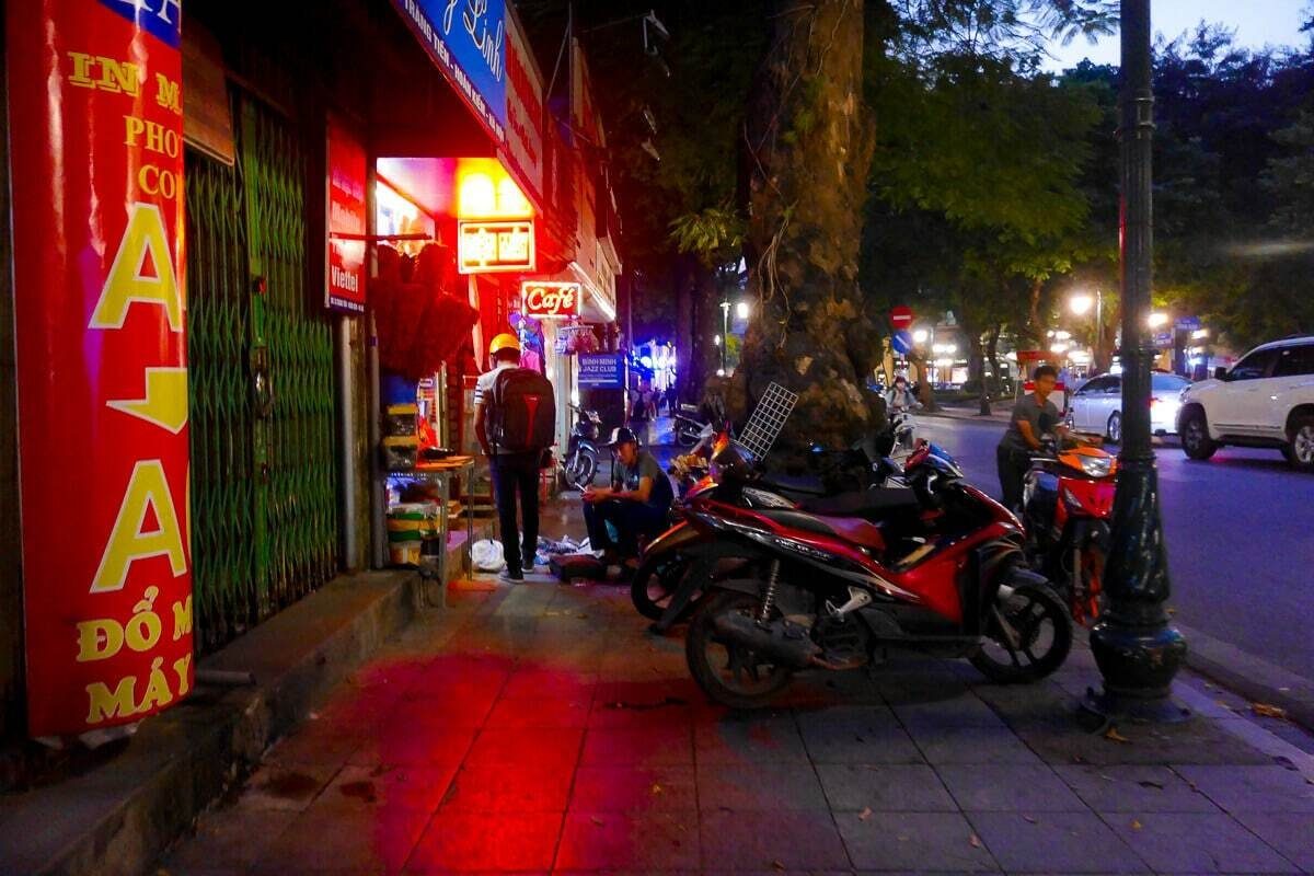 Hanoi sidewalk at dusk with red and yellow shop signs glowing and motorbikes parked under a tree